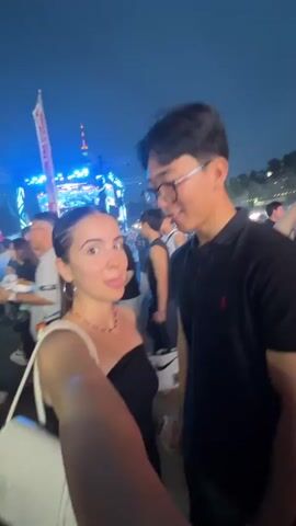 A young woman with long, dark hair pulled into a ponytail and a man with short dark hair and glasses are taking a selfie at an outdoor event at night. The woman is wearing a black off-the-shoulder top and a red beaded necklace. She is holding a white purse. The man is wearing a black polo shirt. They are both smiling. In the background, there is a large stage with bright lights and a crowd of people. The atmosphere appears festive.