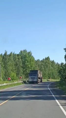 A medium shot captures a gray truck driving down a road bordered by trees. The truck is approaching a curve in the road, indicated by curve warning signs on the left side of the road. The truck continues to turn right, disappearing from view as the camera continues to follow the road.