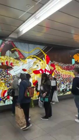 Several individuals are standing and interacting in what appears to be a subway station or underground walkway.  Behind them is a large, vibrant mural depicting a dragon in bright colors. The mural seems to be made of many small pieces, perhaps tiles or mosaics.  The individuals are casually dressed. Some are looking at the mural, while others seem to be talking among themselves.  There is a door to the left of the mural. The location is brightly lit with fluorescent ceiling lights.