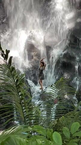 A video captures a woman standing gracefully in front of a powerful waterfall in Gianyar, Indonesia. The woman is positioned on dark rocks, partially submerged in the cascading water, wearing a grey dress. She poses with her arms raised above her head, then lowers them to adjust her hair. The background is dominated by the majestic waterfall, with bright white water rushing over dark, rugged rocks. Lush green foliage, resembling ferns and palm leaves, frames the bottom and sides of the video, creating a natural, tropical foreground. The scene is well-lit by natural daylight, suggesting it is daytime. The prominent sound throughout the video is the immersive roar of the waterfall. The overall impression is one of natural beauty and tranquility.