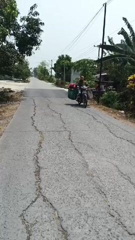 A video captures a person riding a scooter down a cracked asphalt road. The rider, wearing a headscarf, is positioned on the scooter. A large green basket, filled with red objects, is attached to the side of the scooter. The scooter has a dashboard with a speedometer. The road is lined with dry grass and vegetation on one side and trees and houses on the other. Overhead power lines are visible. The sky is bright and clear, suggesting daylight. The video appears to be shot from the perspective of someone riding another vehicle or is a stationary shot of the scene.