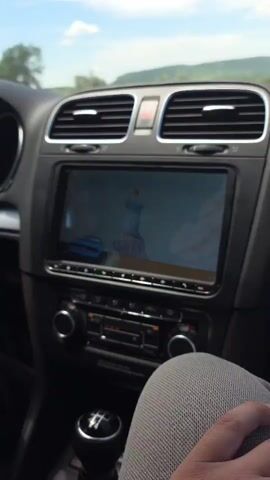 The video shows the interior of a car driving on a highway. The infotainment screen displays an animated cartoon. The car's dashboard and air vents are visible. Outside the car, the road stretches through a green landscape under a blue sky with clouds. The camera is static and focuses on the road ahead.
