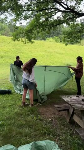 Three people are in a grassy area, seemingly setting up a large green tent. The person in the foreground is wearing denim shorts and a light-colored hoodie, holding a section of the tent fabric. To their left, another person in a dark shirt and shorts is partially visible. On the right, a third person in a brown jacket is also holding the tent. In the background, a green hill rises, covered in grass, and trees line the upper edge of the frame. A wooden picnic table is visible in the lower right corner. Various tent poles and other tent parts are scattered on the ground around the area. The overall environment appears to be outdoors, possibly a park or campsite.