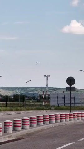 The video shows an outdoor scene at what appears to be an airport or airfield. In the foreground, a row of concrete bollards with red and white stripes lines the edge of a road or tarmac. Beyond the bollards, a chain-link fence separates the road from a grassy area.

In the middle ground, several airport-related structures are visible. There are streetlights, and a tall, lattice tower with antennas. Further back, a large, rectangular building with a white facade is present, also secured by a fence. Another red and white striped tower, possibly a navigation aid or communication tower, stands near this building.

Dominating the sky is a commercial airplane, flying at a moderate altitude. The plane is white with visible engines and wings. The sky is partly cloudy, suggesting daytime with possibly fair weather. The background features rolling hills, covered in green vegetation, hinting at a rural or semi-rural setting surrounding the airport.

The overall impression is a functional aviation environment under a bright sky, with an aircraft in flight as the central focus of activity. There is no visible text in the video.