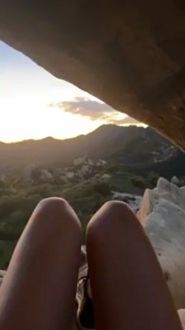 The camera angle is from the perspective of a person sitting with their knees up, overlooking a mountainous landscape at sunset. The person is wearing shoes and their legs are tanned. A large rock overhangs the view, partially obscuring the sky. In the distance, mountains are visible, and the landscape below is covered in green vegetation and scattered rock formations. The sky is a blend of light orange and blue as the sun sets.