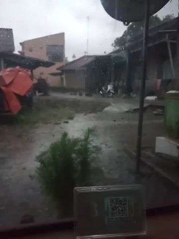Here is a description of the video:

The video is taken from indoors, looking out onto a street during a rain shower. The foreground shows a window with water droplets on it, obscuring the view somewhat. To the right of the window, a clear plastic case displays a QR code. In the background, houses line the street, and a motorcycle is parked near one of the buildings. An orange tarp or covering is visible in the lower left, adding a touch of color to the predominantly gray and muted scene.
