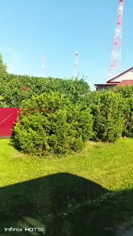 The video shows a view from a vehicle passing by residential properties on a sunny day.  The scene includes green lawns, trees and bushes, houses with red roofs, a garage with a red door, a white minivan parked in a driveway, and several tall communication towers in the background.  The camera is moving, indicating the perspective of someone in a moving vehicle.  The ground appears to be a mix of grass and some patchy areas. A red fence is visible in several shots. There are no people visible in the video.