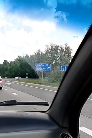 The video shows a view from inside a car driving on a highway. The dashboard is visible in the foreground. Outside the car, there are trees lining the highway, a blue sky with white clouds, and other cars on the road. One car is in front of the vehicle, and several others are in the distance. A road sign is visible on the right side of the road. The camera is angled upwards, showing the windshield.