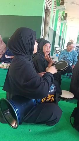 The video shows a group of people sitting on a green carpet, likely indoors or under a covered outdoor area. In the foreground, a woman wearing a black hijab and black clothing is playing a dark blue goblet drum positioned between her legs. She is facing to the right of the frame and her hands are visible, appearing to strike the drum. Next to her, also wearing a black hijab and black clothing, is another woman who is clapping her hands. Behind these two women, to the right, a man wearing a blue shirt and a white cap is sitting and playing a black drum. In the background, another person wearing a light pink hijab is seated on the left side of the frame. The environment appears to be a simple room with green walls and a white ceiling with exposed beams.