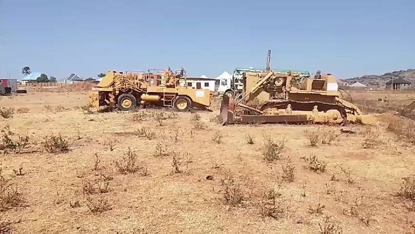 The video depicts two pieces of heavy machinery, a scraper and a bulldozer, sitting idle on a dry, grassy field. The scraper, painted yellow, has a large hopper and multiple wheels. The bulldozer, also yellow, features a prominent blade at the front and tracks for movement. In the background, a white truck, buildings, and low-lying hills can be seen. The sky is clear and blue, indicating a sunny day. Small, brown bushes are scattered across the foreground. A fence and some huts can be seen in the distance to the right.