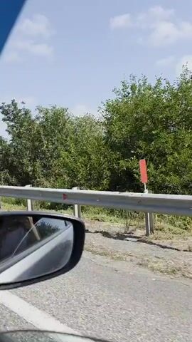 The video shows the interior of a car driving on a highway. The camera is positioned to capture the road ahead, the side mirror, and the passenger side window. Through the side mirror, a person can be seen in the passenger seat, looking down. Outside the car, the highway is visible with other vehicles, including cars and trucks. Mountains and trees line the side of the road, under a clear blue sky with some clouds. The video appears to be recorded during the daytime.