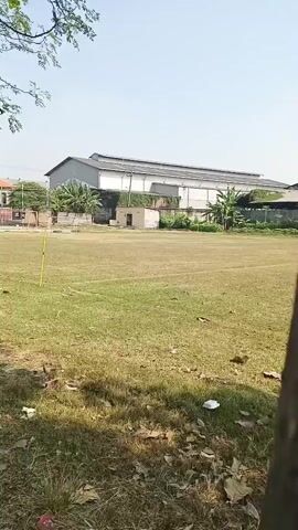 The video shows a wide shot of an open field with dry grass. There is a large, industrial-looking building in the background, with a white roof and grey walls. Several trees line the left side of the frame, casting shadows on the ground. In the middle ground, several children are visible playing on the field. The children are scattered across the field and appear to be running and playing games. There are also several black tires stacked on the ground near the edge of the field. The sky is clear and blue, indicating a sunny day. The overall impression is of a somewhat neglected outdoor space being used for recreation by children.