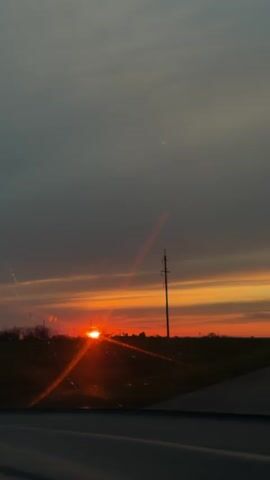 The video captures a stunning sunset viewed from a moving vehicle in what appears to be a rural or semi-rural setting. The sky is dominated by deep gray and bluish clouds at the top, gradually transitioning into vibrant horizontal bands of orange, yellow, and red hues as it approaches the horizon. The sun, a bright orange disc, is partially visible just above the horizon, casting a warm glow and light flares onto the vehicle's windshield. A tall utility pole stands prominently in silhouette against the colorful sky on the right side of the frame. A dark asphalt road is visible in the foreground, with faint outlines of trees or bushes in the distance along the horizon. The time of day is clearly sunset, and the weather appears partly cloudy with clear visibility towards the horizon. No text is visible in the video.