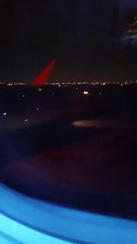 The video shows a view from an airplane window at night.  The wing of the plane is visible in the foreground, and the lights of a city stretch across the horizon. There is a light on the ground closer to the plane. The video is dark overall.