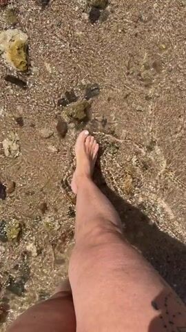 A person walks barefoot on a beach. The sand is a mix of tan and white, interspersed with small rocks and what appear to be pieces of coral. Clear water washes over the sand and around the person's foot as they walk. The scene is brightly lit by sunlight.