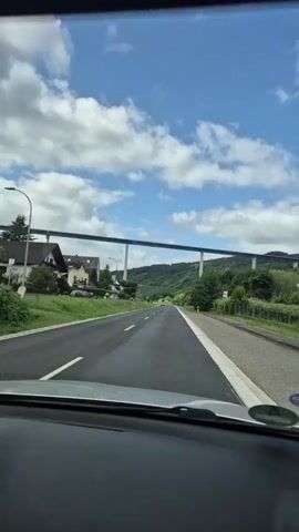 A first-person view from inside a moving car shows a two-lane asphalt road with a solid white line on the right shoulder and a dashed white line separating the two lanes. A large bridge crosses the road horizontally in the distance. On the left side of the road, there are houses and green vegetation. On the right, there are trees and a slight incline of a hill. The sky is partly cloudy with blue patches visible. The car's dashboard and windshield are visible in the foreground.