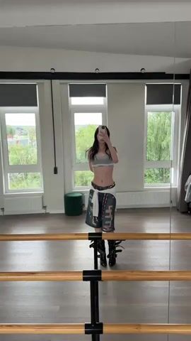 A young woman with long dark hair, wearing a gray Calvin Klein sports bra and ripped denim shorts, stands in a dance studio. She is taking a selfie in a large mirror. The studio has large windows overlooking a green, tree-filled landscape. There is a barre in the foreground of the mirror's reflection, and a green stool is visible to the left of the woman. The woman moves her arms and head slightly as she records the video.
