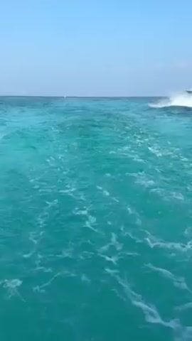 A boat with several people onboard is visible in the distance, moving across a body of water with a turquoise color. The camera appears to be positioned on another boat, as evidenced by the churning water and wake stretching behind. Above, a clear blue sky is sparsely dotted with a few small clouds.