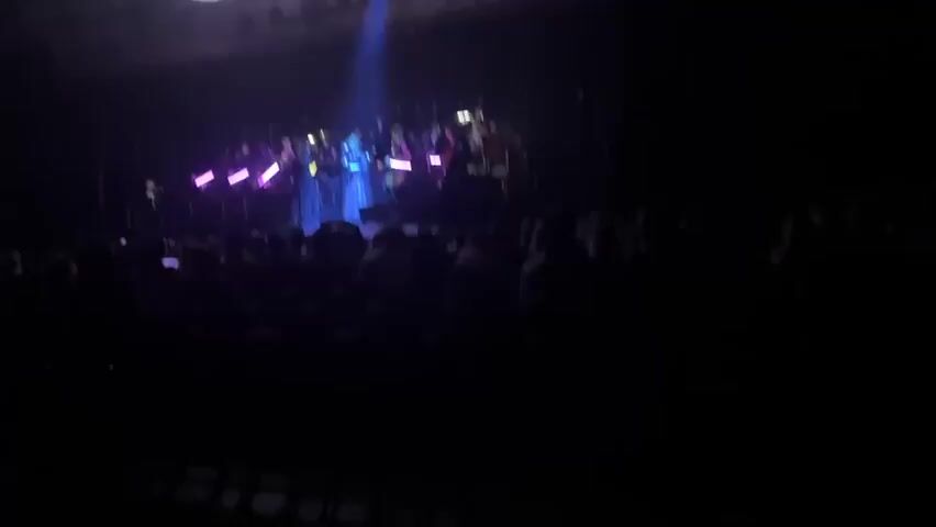 An orchestra is performing on a stage in front of a seated audience. The stage is dimly lit, with a blue spotlight shining on a woman in a blue gown singing into a microphone. The other members of the orchestra are playing their instruments, including violins, cellos, and percussion. The violins have purple light sticks attached. There is a large, circular object hanging above the stage. The audience is mostly silhouetted.
