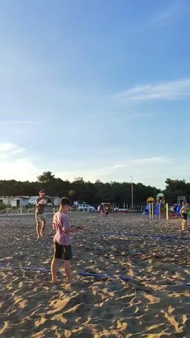 A person is serving a volleyball on a sandy beach court. The server is a young male wearing a purple t-shirt and dark shorts. He is looking at the ball, which he holds in his hands. He is in the process of tossing the ball up to serve it. The court is marked with blue lines. In the background, another young male is standing with his hands in his pockets, watching the server. He is wearing a gray shirt and shorts. Beyond the court, there are some trees and a few buildings. The sky is clear and blue. The sun appears to be setting, casting a warm glow on the scene.