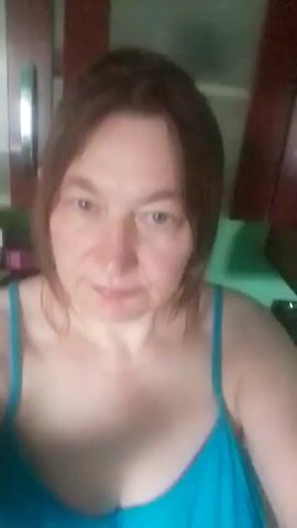 A medium shot video shows a woman with shoulder-length brown hair and light skin facing the camera. She is wearing a teal-colored tank top with thin straps. The background appears to be a kitchen, with dark wood cabinets above and a green surface below. The lighting is soft and appears to be indoor lighting. The woman's expression is neutral, and she is not speaking. The video is short, lasting only a few seconds, and there are no discernible actions or interactions occurring within the frame. The focus is on the woman's face and upper body.