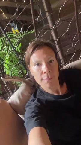 A mid-shot captures a woman with shoulder-length brown hair, wearing a black t-shirt, as she sits comfortably in a cushioned chair. The chair, made of a lattice-like metal frame, is positioned outdoors, with lush green ferns visible behind it. The background includes glimpses of the surroundings, with the presence of string lights adding a soft glow. The woman's gaze is directed toward the camera, and she remains still throughout the video.