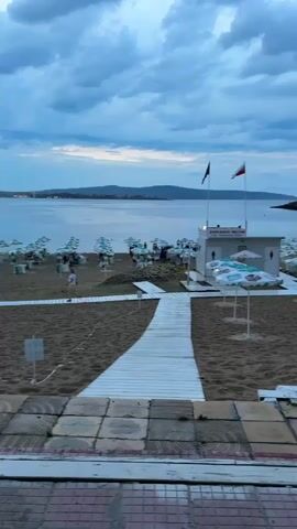 The video shows a beach scene from an elevated perspective looking towards the sea. The sky is overcast with dramatic clouds. On the beach, there are many rows of beach umbrellas, most of which are white with teal accents. Some lounge chairs are visible under the umbrellas. A few people are scattered across the beach, some walking and some sitting. There is a white building with two flags flying above it, one black and one Bulgarian flag. A white wooden walkway extends from the foreground into the sand, creating a path towards the beach. The sea is calm, and distant hills are visible on the horizon. The foreground shows a tiled surface. The overall mood appears to be transitioning from day to evening, with muted light.