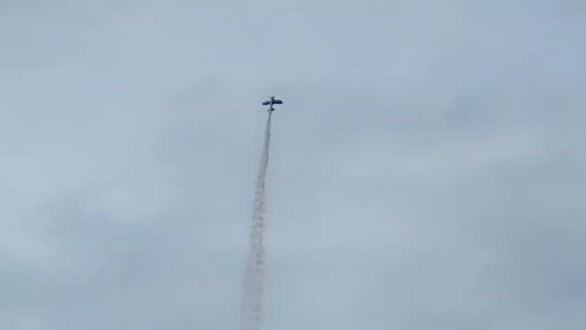 A small, dark airplane is seen performing an aerobatic maneuver against a cloudy, gray sky. The plane is ascending vertically, leaving a thick trail of brown smoke behind it. The smoke plume starts wide and narrows as it ascends, indicating the plane's rapid ascent. The plane itself is a tiny silhouette against the vast sky. The camera angle is looking up at the plane from below.