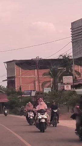 The video shows a road in what seems to be a tropical area. The sky is overcast and has a reddish-pink hue, suggesting it may be near sunrise or sunset. There are several individuals riding motorbikes along the road.  Some of the riders are wearing headscarves. There is a building under construction, appearing to be brick, with a partially covered roof. There are other structures visible, including houses with corrugated metal roofs. Utility poles and power lines are present overhead. The road itself is paved with a yellow line marking the center.  There are trees and foliage along the roadside.