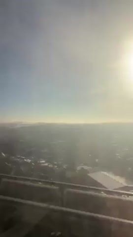 The video shows a view of a city from a high vantage point, likely a building or tower. The sky is clear blue with a bright sun shining through. The city below is covered in a layer of haze or smog, obscuring details, but buildings and trees can be seen. A railing is visible at the bottom of the frame, suggesting the view is from a balcony or window. The camera is still and the focus is on the city and sky.
