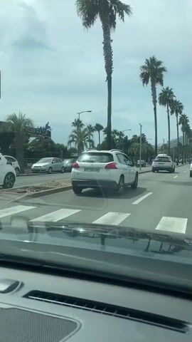 The video shows a view from inside a car, looking out at a street scene with palm trees and several cars. The car appears to be moving slowly or stopped in traffic. On the left side of the road, there is a building with a sign that says "CLAIRAZUR". Several cars are parked or driving on the street. The sky is cloudy. The interior of the car, including part of the dashboard and windshield, is visible in the foreground. The video is shaky, suggesting it was filmed while the car was in motion.