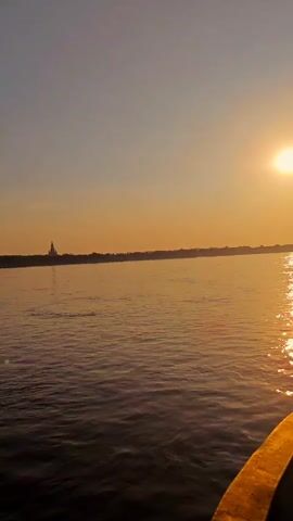 A video taken from a boat on a body of water, possibly a river. The sun is setting on the right side of the frame, casting a warm, orange glow across the sky and reflecting on the water. A silhouette of a distant shoreline with some structures, possibly buildings or trees, is visible on the left side. The front right edge of the boat is visible in the lower right corner of the frame. The water appears calm with gentle ripples. The overall mood is serene and peaceful.