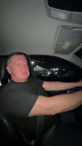 A person is sitting in the driver's seat of a car at night, holding the steering wheel with both hands. They are wearing a black t-shirt and dark pants. The car's interior is visible, including the dashboard and a sun visor. Outside the car, streetlights and distant buildings can be seen. The camera appears to be positioned inside the car, possibly on the passenger side, filming the driver from the side.