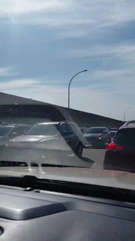 The video shows a view from inside a vehicle looking forward through the windshield. The vehicle is in traffic with other cars around. In the middle distance, a person on a motorcycle is weaving through the stopped or slow-moving vehicles. The motorcycle rider is wearing a helmet and dark clothing. The weather appears to be sunny with some clouds in the sky.