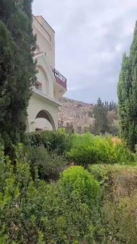 The video pans across a courtyard with a walkway lined by manicured hedges and cypress trees. A building with arched entrances is visible on the left, and a domed building with an orange roof and windows is seen on the right. The sky is overcast.