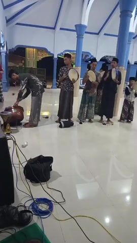 The video shows a group of young males in a large, indoor space with a high ceiling supported by blue columns. The floor is reflective and appears to be tile. In the foreground, there are several audio cables and a black bag, suggesting some kind of sound equipment is present. In the background, a group of five young males are standing in a line. One male on the far left is bent over, appearing to adjust a large drum. The other four males are holding what appear to be tambourines or frame drums. They are dressed in traditional Islamic attire, including sarongs and skullcaps. The atmosphere suggests a musical performance or practice is underway. The lighting is artificial and casts some shadows. The overall environment seems to be a mosque or similar religious building.