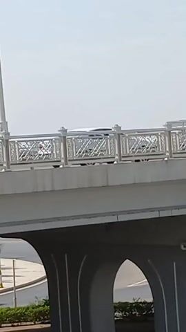 The video shows a close-up of a bridge. The bridge has a light-colored railing with an ornate, geometric design. Below the bridge, there are arched structures. The sky is bright and clear, suggesting it is daytime.