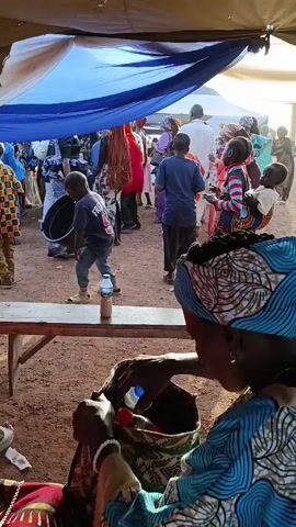 The video shows a gathering of people under a tent, with a dirt ground visible. Several children are running around, and many adults are standing and talking. A woman wearing a colorful headwrap and a blue patterned shirt is sitting near the camera, seemingly occupied with something in a bag. A table stands in front of her with a bottle of water placed on it. In the background, there's a mix of colorful clothing, suggesting a lively event or celebration. The sunlight filters through the tent covering, creating a bright and open atmosphere. A man dressed in a white outfit is walking by. The scene is filled with activity and people, suggesting a social gathering.