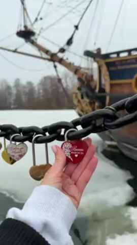 A hand holds a red, heart-shaped padlock with the words "Vmecte Navsegda" written in Cyrillic script, attached to a chain with other locks. The chain is likely part of a bridge or barrier, and in the background, there is a body of water partially covered with ice and a large, old-fashioned ship. The sky is overcast.