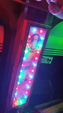 The video shows a close-up, angled view of a long, rectangular wooden box adorned with colorful flashing LED lights. Inside the box, a framed picture is displayed, featuring what appear to be religious figures. The top part of the picture shows an elder man with a long white beard and a turban, surrounded by smaller circular images. Below this, there's an image of a large building, possibly a temple. Further down, two more figures are visible; one man with a beard and a turban holding a bird, and another man in a turban. The box itself is red and has a decorative golden frame around the picture. The background is dark, with hints of green and red lighting. The video appears to be taken in a dimly lit environment, possibly at night.