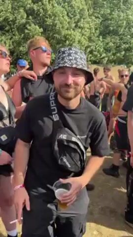 Here's a description of the video:

The video captures a man at an outdoor event, likely a music festival, on a sunny day. He's in the foreground, directly facing the camera, wearing a black T-shirt, black pants, and a distinctive bucket hat with the word "CUNT" printed repeatedly on it. He also has a black Supreme-branded fanny pack slung across his chest. He holds a plastic cup containing a light-colored beverage, possibly beer, in one hand and a lit cigarette in the other.

The man seems to be enjoying himself, offering a smile as he looks at the camera and gestures with his hands. Behind him, there's a crowd of people, some wearing festival attire, and green foliage suggesting a wooded or park-like setting. The atmosphere appears lively and festive, with people moving around and socializing. Music can be heard in the background.