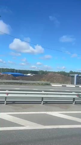 The video begins with a view of a bus stop with a sign that appears to be in Russian. The sky is blue with scattered white clouds. In the background, there is a large pile of dirt and debris, suggesting a construction or excavation site. Various construction vehicles, including an excavator, can be seen in the distance. A speed limit sign indicating 70 is visible along with a road construction warning sign. The road in the foreground has white lane markings and a metal guardrail. The scene appears to be in a rural or semi-rural area with trees forming a distant treeline. The camera is moving, likely from a vehicle, as the perspective shifts slightly throughout the short clip.