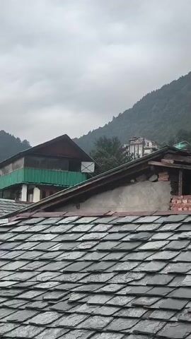 THE VIDEO SHOWS A CLOSE-UP VIEW OF A SLATE ROOF IN THE FOREGROUND. BEYOND THE ROOF, THERE ARE SEVERAL BUILDINGS AND A WOODED MOUNTAIN IN THE BACKGROUND. ONE OF THE BUILDINGS IS A MULTI-STORY RESORT WITH THE WORDS "JR RESORT" VISIBLE ON THE FACADE. THE SKY IS OVERCAST.