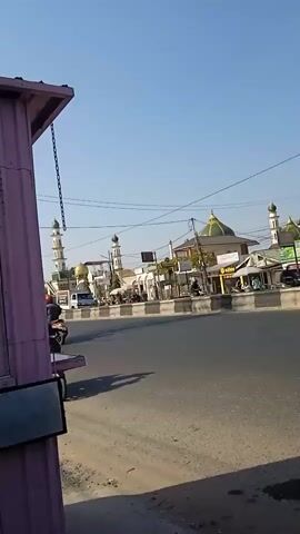 The video shows a street scene with a mosque in the background. In the foreground, two motorcycles are visible on the road. The first motorcycle, a red scooter, is ridden by a woman wearing a floral dress, with a child sitting behind her. The second motorcycle is a white scooter carrying two people. The sky is clear and blue, indicating a sunny day. The overall environment appears to be an urban or semi-urban area, possibly in a region with Islamic cultural influence due to the prominent mosque.