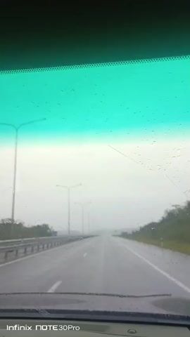 The video captures a view through a car windshield, showing a wet, empty highway under a cloudy sky. Raindrops are visible on the windshield, distorting the view slightly. The road ahead is wet and appears to be a highway with streetlights and guardrails on either side. The sky is overcast and the overall atmosphere is gloomy, suggesting it is raining or has recently rained. There are no other vehicles or people visible on the road. A watermark at the bottom of the screen indicates the video was recorded with an Infinix NOTE 30Pro.