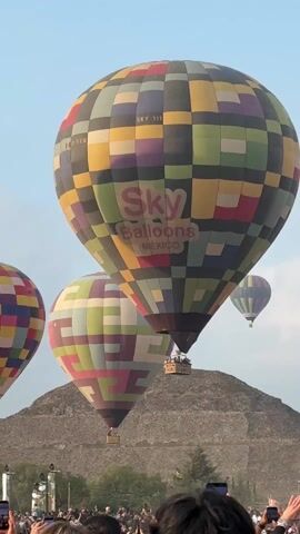 The video shows four hot air balloons, operated by "Sky Balloons MEXICO," in the air against a mostly clear, blue sky. The balloons are patterned with colorful squares. In the background is a large pyramid-like structure. At the bottom of the video, a crowd of people can be seen holding up their phones, seemingly recording the event. A black metal structure is visible on the left side of the frame. Music is playing in the background.