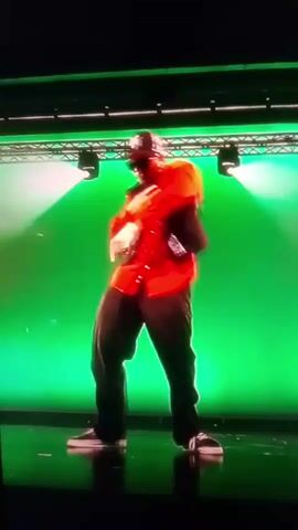 The video shows a person dancing in front of a green backdrop. The person is wearing a red shirt, a black vest, black pants, a baseball cap, and sunglasses. They are performing dance moves with their arms and legs. The lighting is bright, with spotlights illuminating the stage. The video is recorded from a low angle, looking up at the dancer.