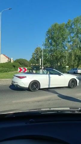 This is a dashcam recording from a vehicle driving on a sunny day in Písek, Czechia. The scene is an intersection with a roundabout. A white convertible car with a person in the passenger seat drives past, followed by a grey sedan and then another grey sedan. In the background, there are trees, bushes, a building with a red roof, and streetlights. A road sign with two red and white arrows indicating a curve to the right is visible. The weather appears to be clear and sunny.