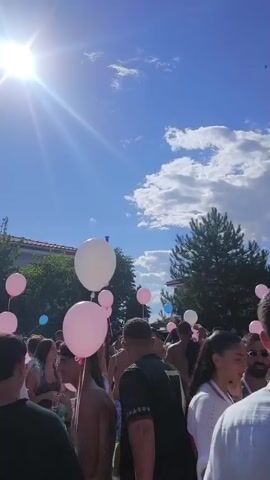 The video shows a group of young adults outdoors on a sunny day. They are gathered in what appears to be a party or celebration, with many holding pink, white, and blue balloons. Some individuals are wearing swimwear. The background includes trees and a building. The sun is prominently visible in the sky, creating a bright glare in some shots. People are interacting with each other, and the atmosphere seems festive.