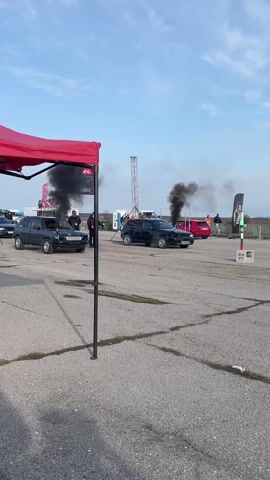 Two gray Volkswagen Golfs are engaged in a drag race on a paved track. Both cars emit thick black smoke as they accelerate, leaving trails of dust. The car on the left is a lighter shade of gray and has a chrome exhaust pipe. Spectators stand behind a barrier to the right. In the background is a rural landscape with a partially cloudy sky. A red canopy and advertising banners are on the left side of the scene. A metal support structure, a traffic light, and more spectators are further down the track.